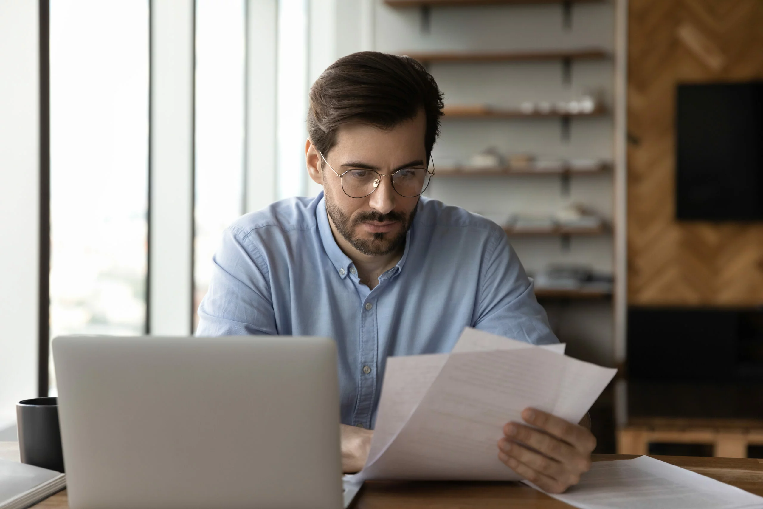 A man with glasses sitting at a desk, surrounded by papers and documents.